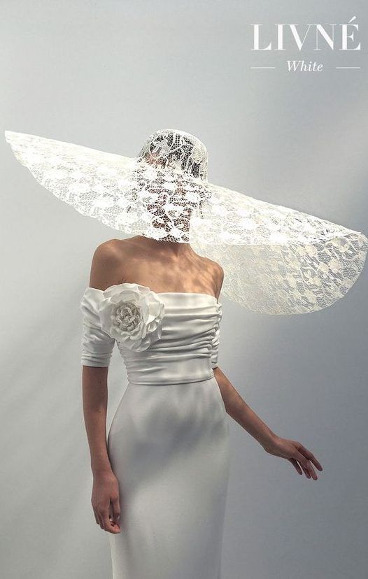 Свадебное платье цвета слоновой кости без рукавов, декольте с открытыми плечами, складчатые детали, крупный цветок из крепа. Шляпка невесты - оригинальное завершение образа. Livné White. Коллекция 2020